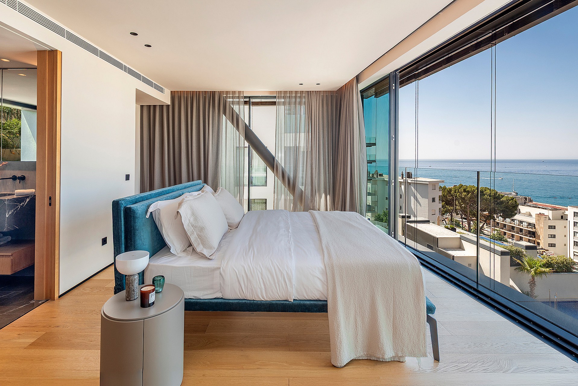 Modern master bedroom with a teal upholstered bed, light oak flooring, and expansive glass walls overlooking the ocean.