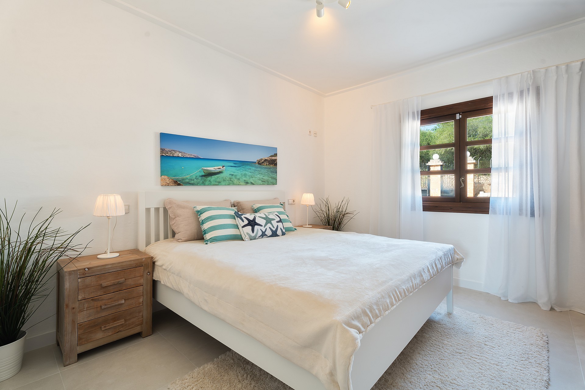 Dormitorio luminoso con cama blanca, cajoneras de madera y contraventanas oscuras con cortinas ligeras.