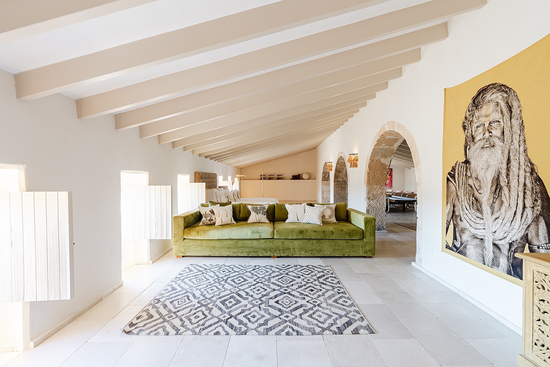 Wide shot of a bright attic living space featuring a long olive green velvet sofa, white tiled floor, and exposed timber beams.