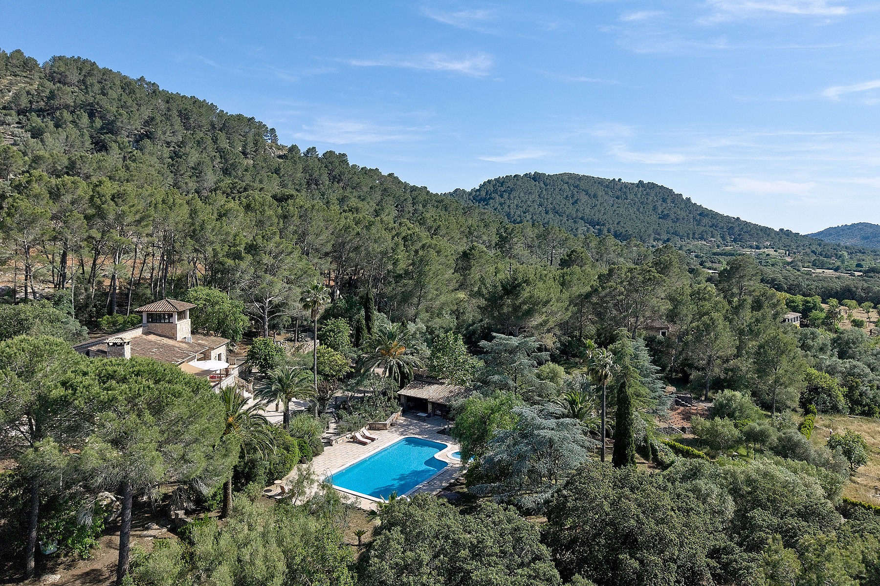Aerial perspective of a stone finca surrounded by pine forests, showing a swimming pool and terraced gardens in Alaro.