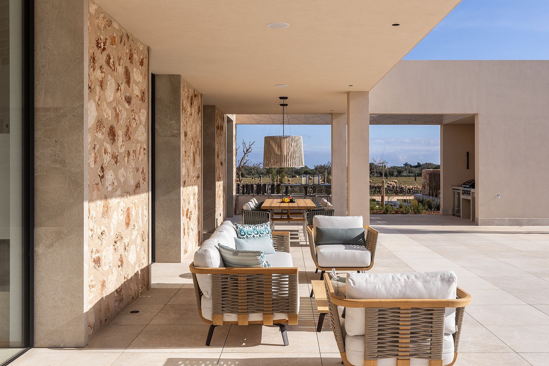 A spacious covered terrace with stone walls, outdoor lounge seating, and a dining area, overlooking a distant mountain landscape.