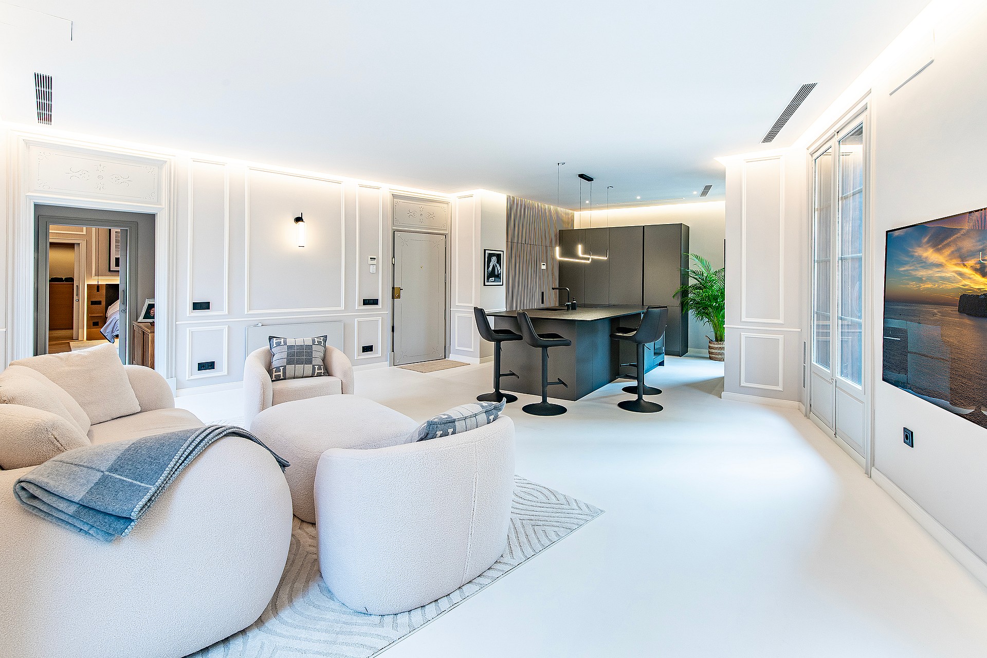 Wide shot of a modern white open-plan living room with a kitchen island, textured armchairs, and floor-to-ceiling windows.