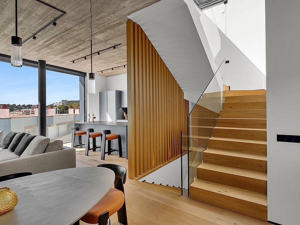 Interior of a modern penthouse featuring a wooden staircase, concrete ceiling, and open-plan kitchen with a breakfast bar.
