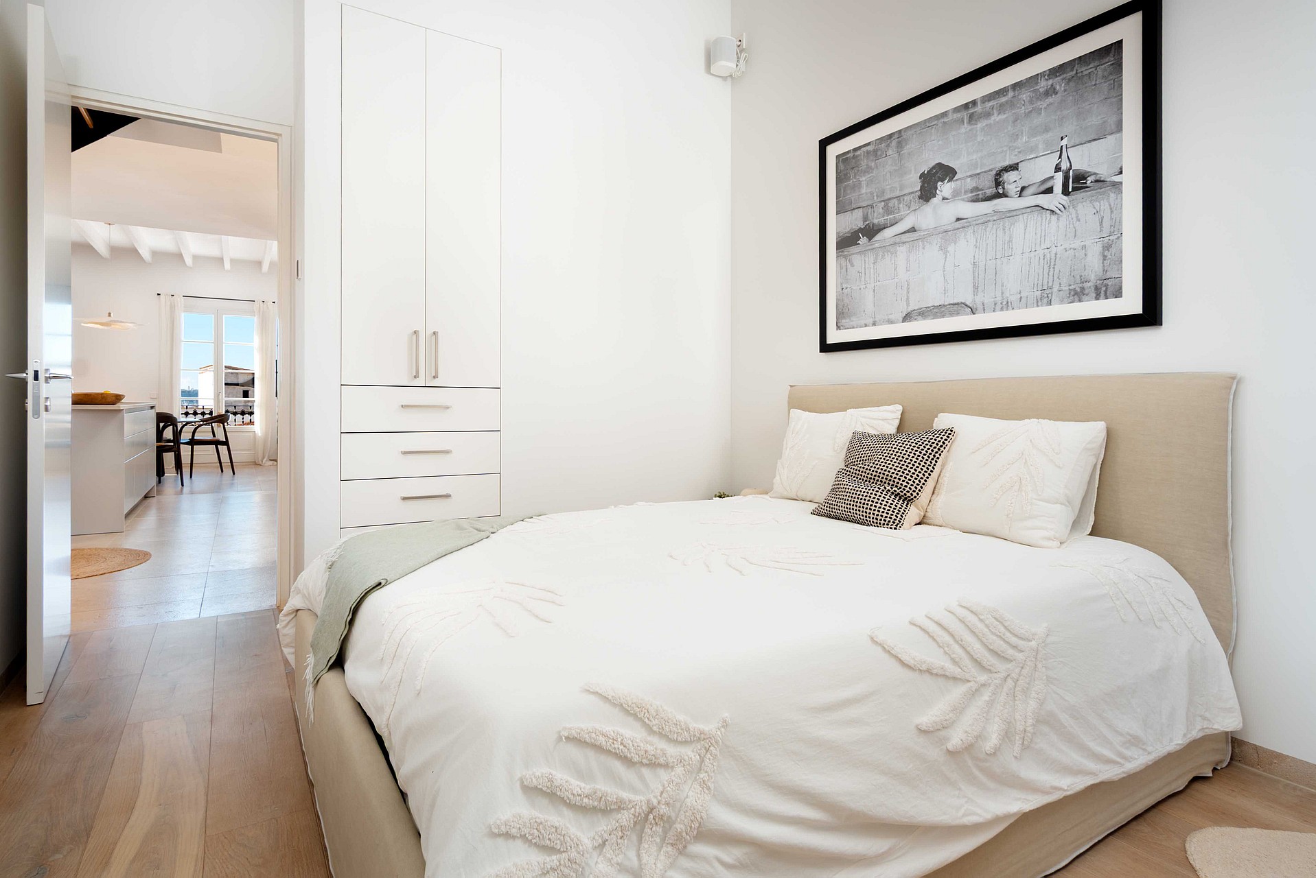 A minimalist bedroom featuring a beige upholstered bed, white textured bedding, and a framed black and white landscape print.