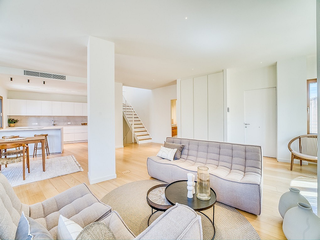 Modern open-plan living room with light oak flooring, grey tufted sofas, and a white architectural staircase.