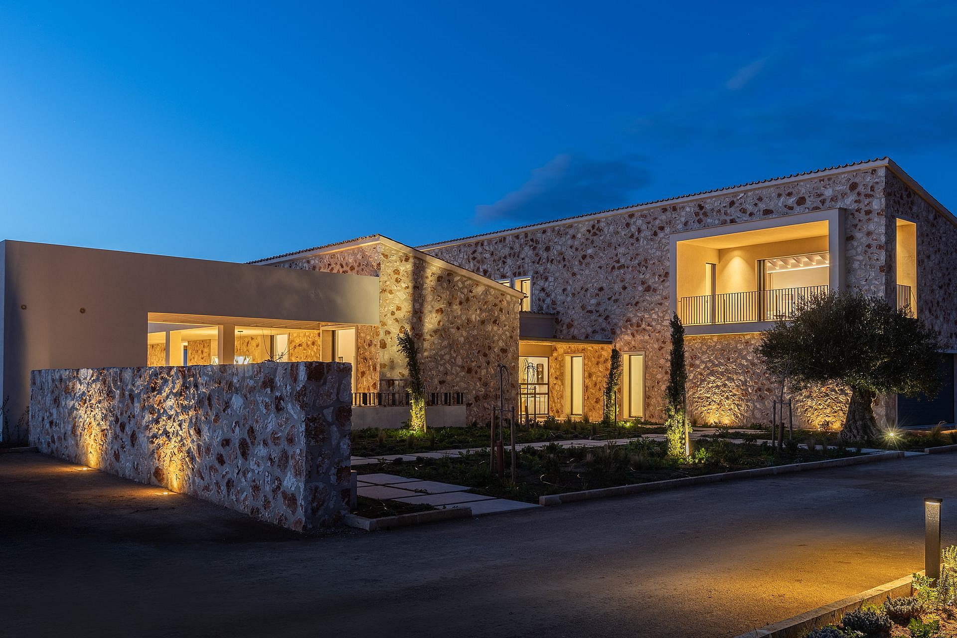 Exterior view of a modern stone-clad finca in Santanyí at twilight, showing illuminated interior spaces and landscaped grounds.