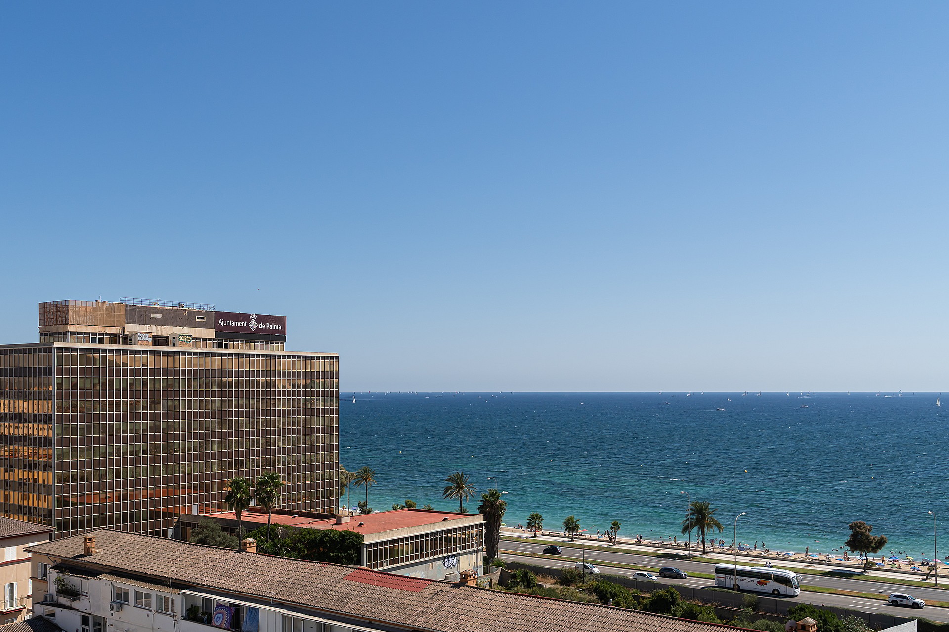 High-angle view overlooking a modern commercial building with mirrored glass facade, a sandy beach, and the Mediterranean Sea.