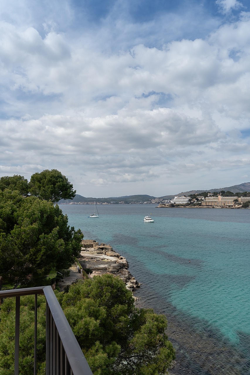 View from a terrace railing over turquoise water, a rocky shoreline, and yachts anchored in a Mediterranean bay.