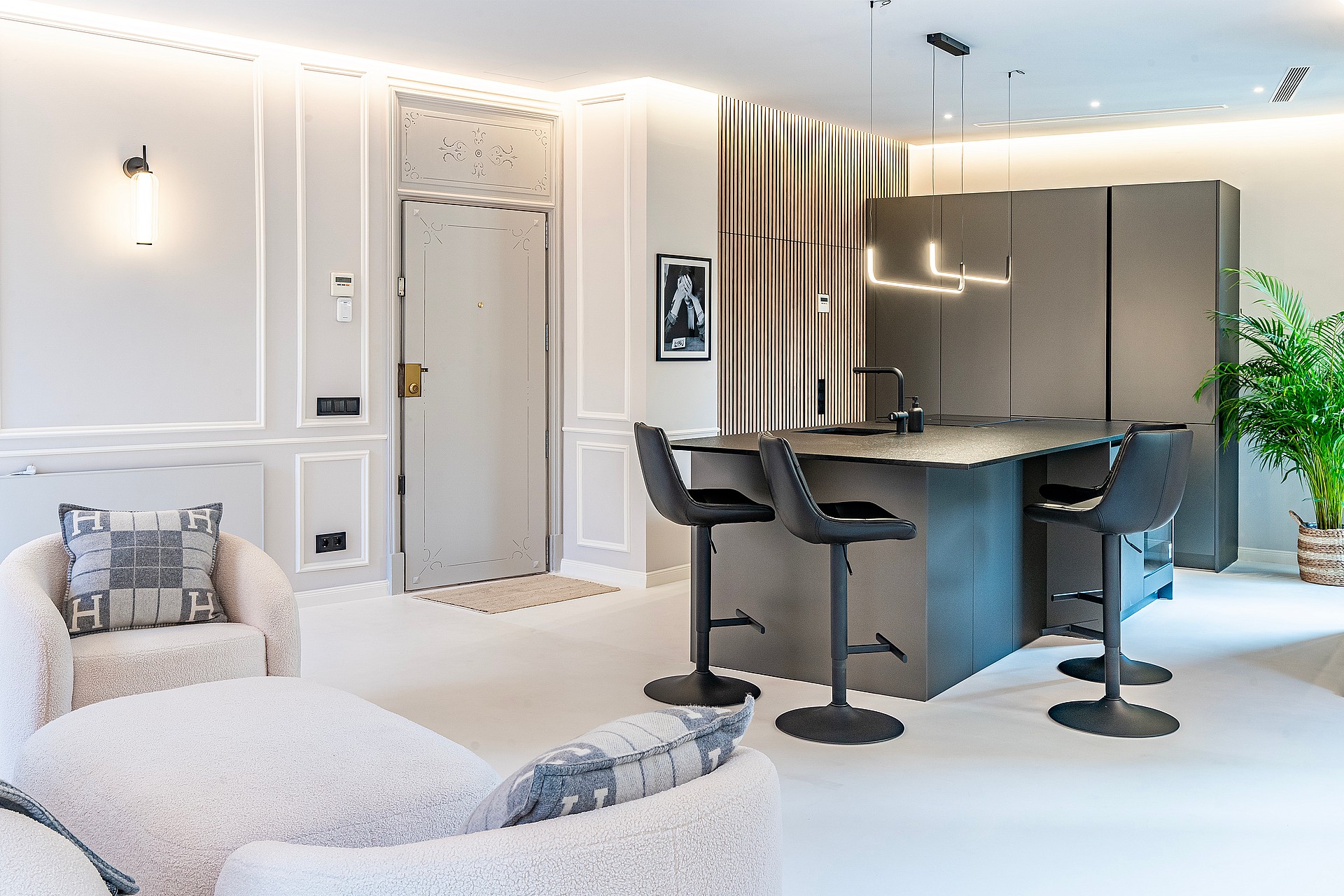 A modern open-plan interior featuring a dark kitchen island with bar stools, neutral textured seating, and wall moldings.
