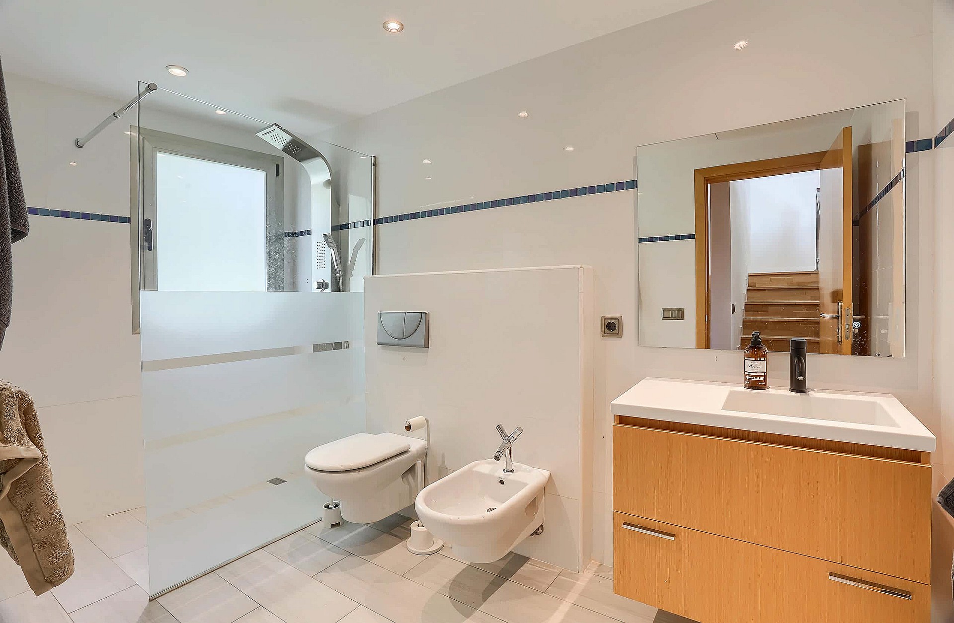 Bright bathroom featuring a walk-in shower with glass partition, wall-hung toilet, bidet, and a wooden vanity unit with mirror.