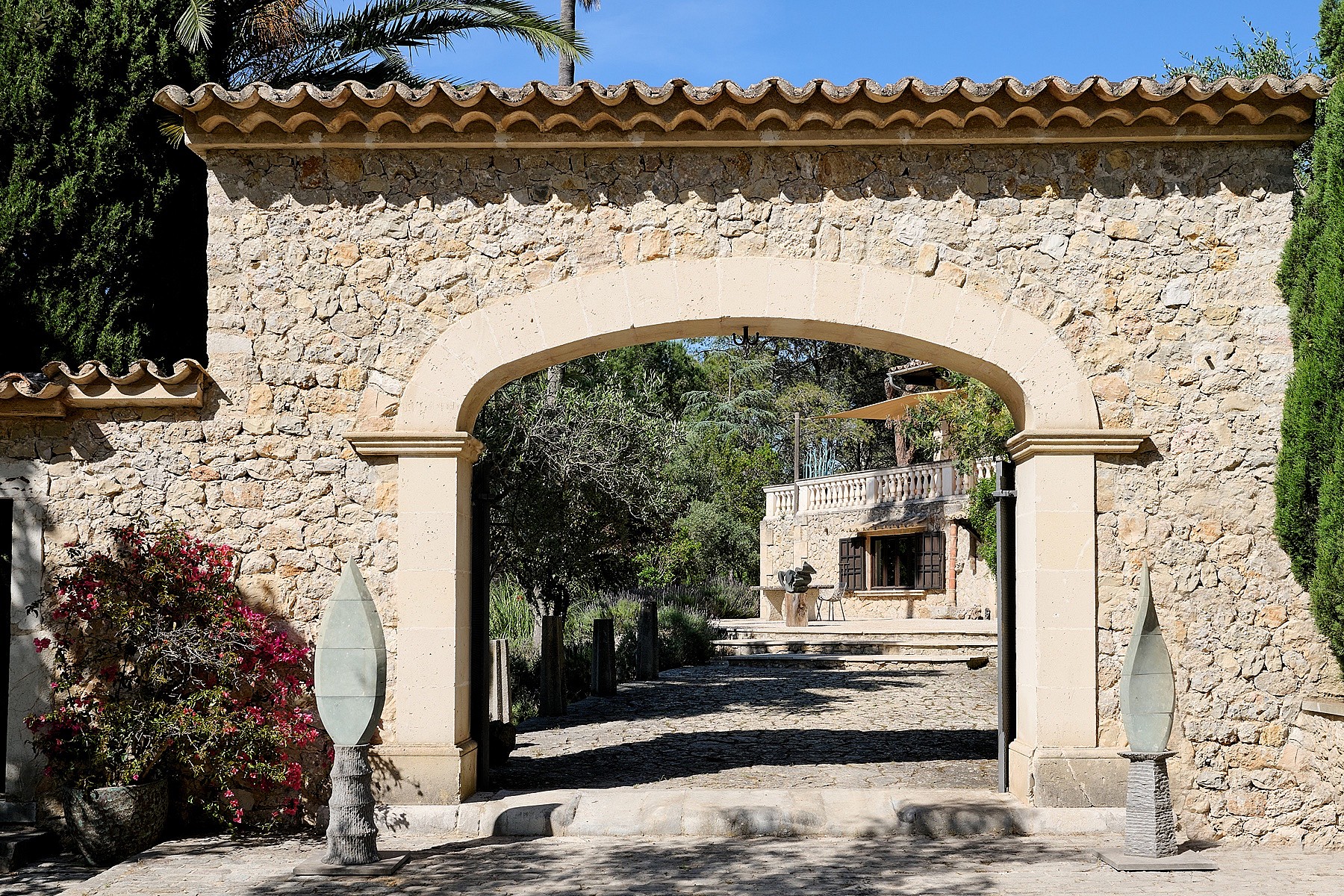 A traditional stone archway entrance to a finca, featuring rough-hewn masonry and clay roof tiles, opening to a gravel path.