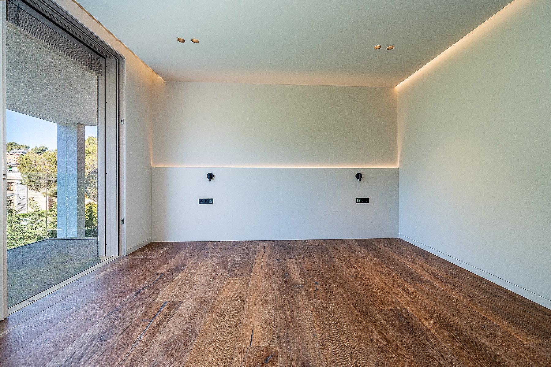 Empty bedroom with light-colored walls, recessed strip lighting, and wide-plank wood flooring opening to a terrace.