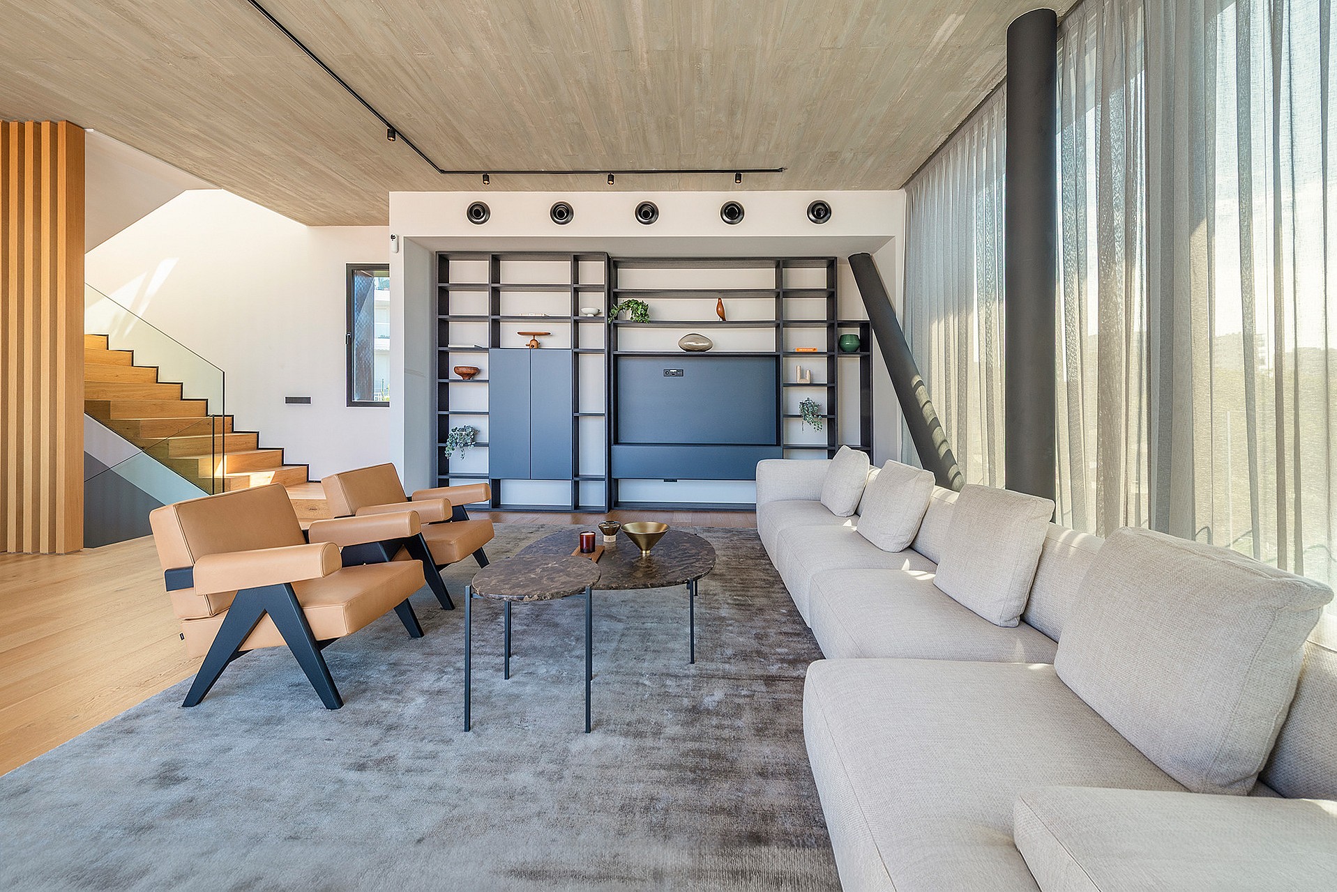 A modern open-plan living room with a textured concrete ceiling, tan leather armchairs, a large grey sofa, and a black metal shelving unit against a white wall. A wooden staircase with glass railings is visible to the left.