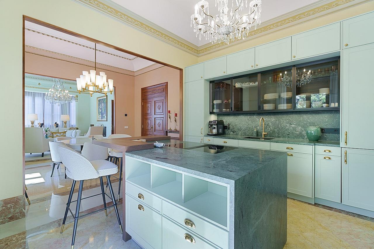 Modern kitchen with sage green cabinets, a dark grey stone island, and bar stools, leading to a dining area with a chandelier.