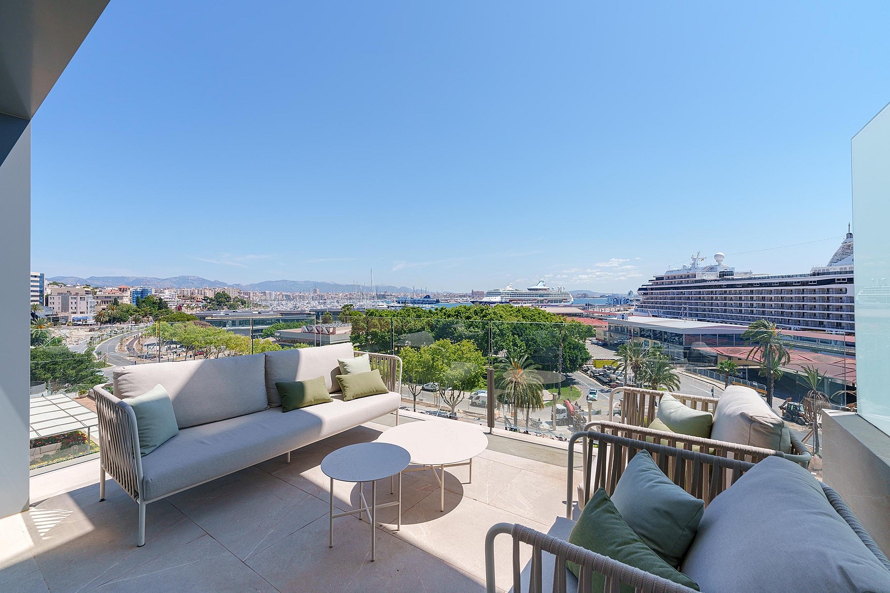 Terraza amplia con sofás grises de exterior y cojines verdes, suelo de piedra y vistas al puerto y al mar.