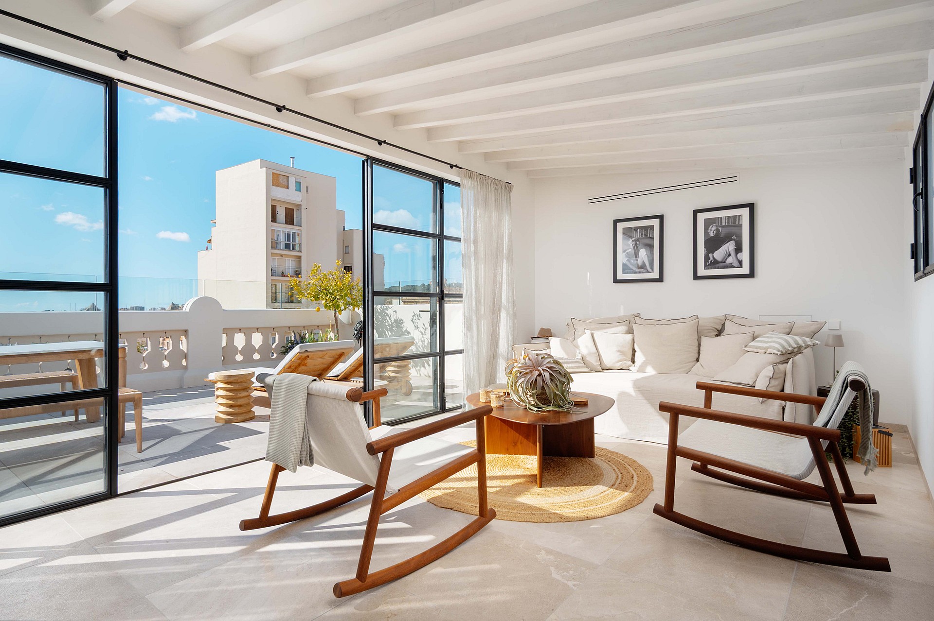 Upper-level sitting room with wooden rocking chairs and a cream sofa, steel-framed doors open to the terrace and rooftops beyond.