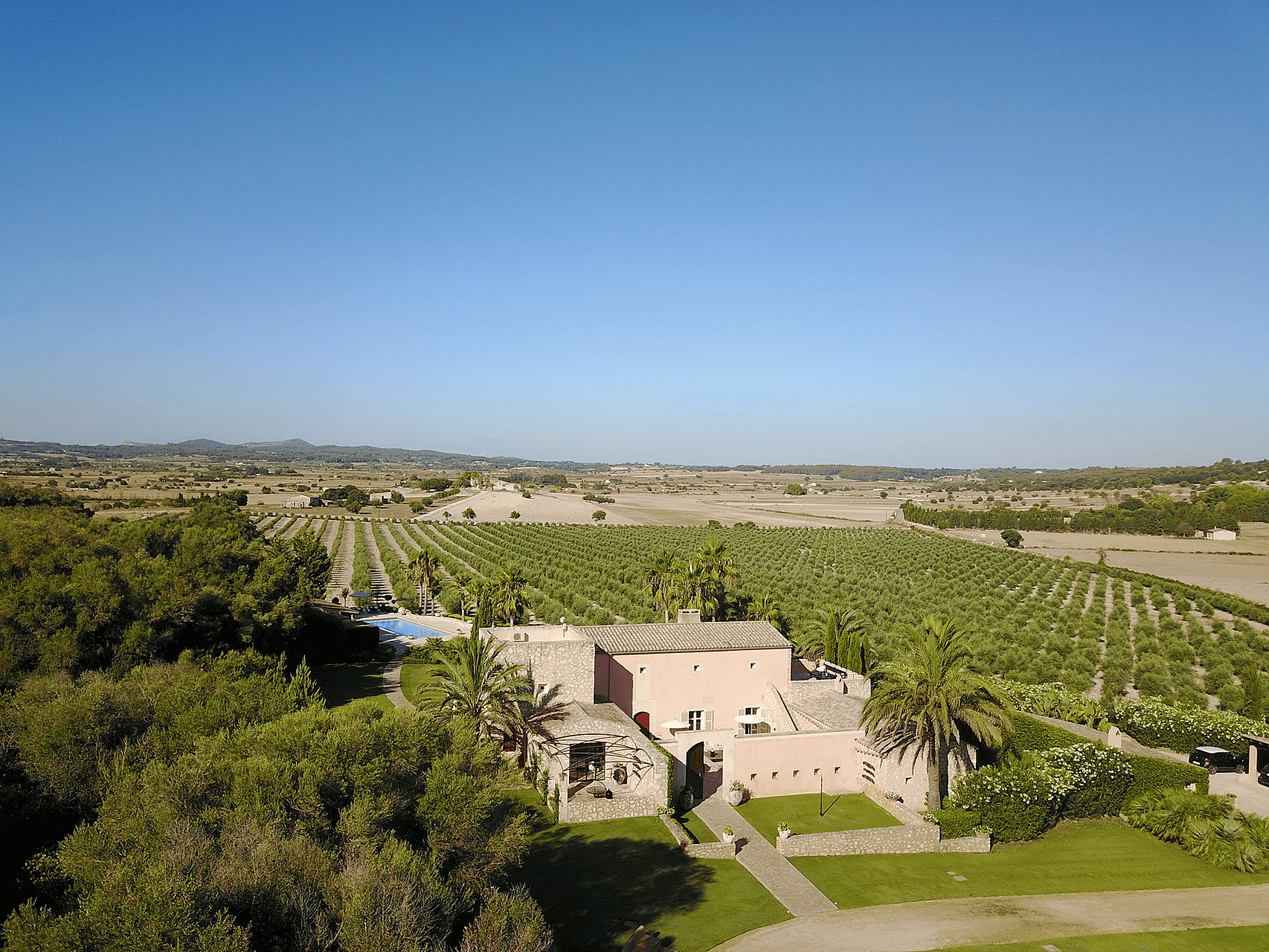 Aerial drone shot of a pink-walled Finca surrounded by vast agricultural fields and Mediterranean landscape.