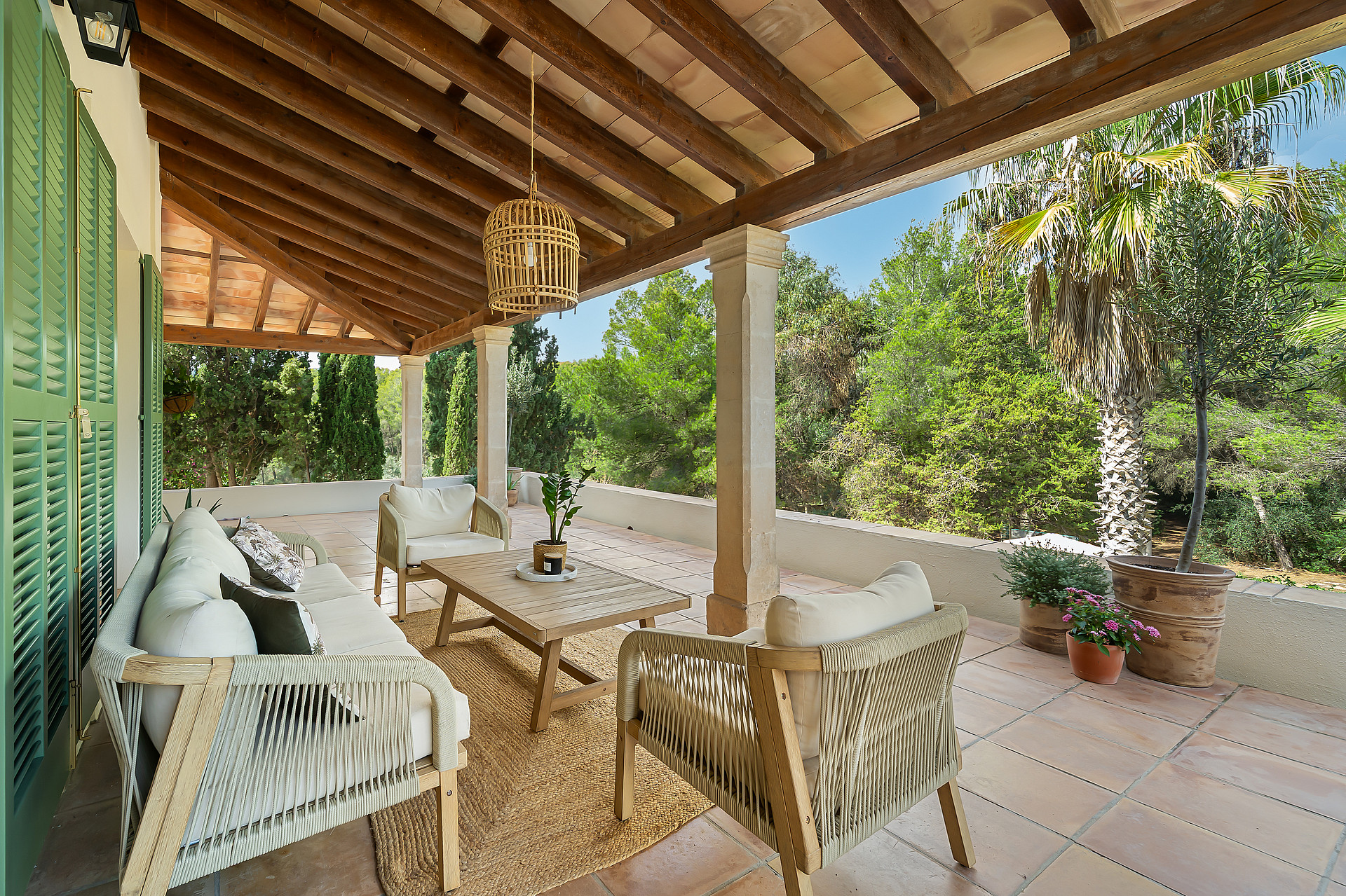 A covered terrace with a timber-beamed ceiling, stone pillars, and neutral lounge furniture overlooking a forest.