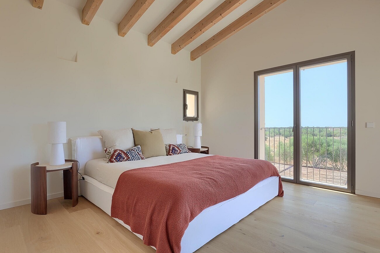 A bedroom with light wood flooring, white walls, and exposed ceiling beams. A large bed with a red cover faces a terrace door.