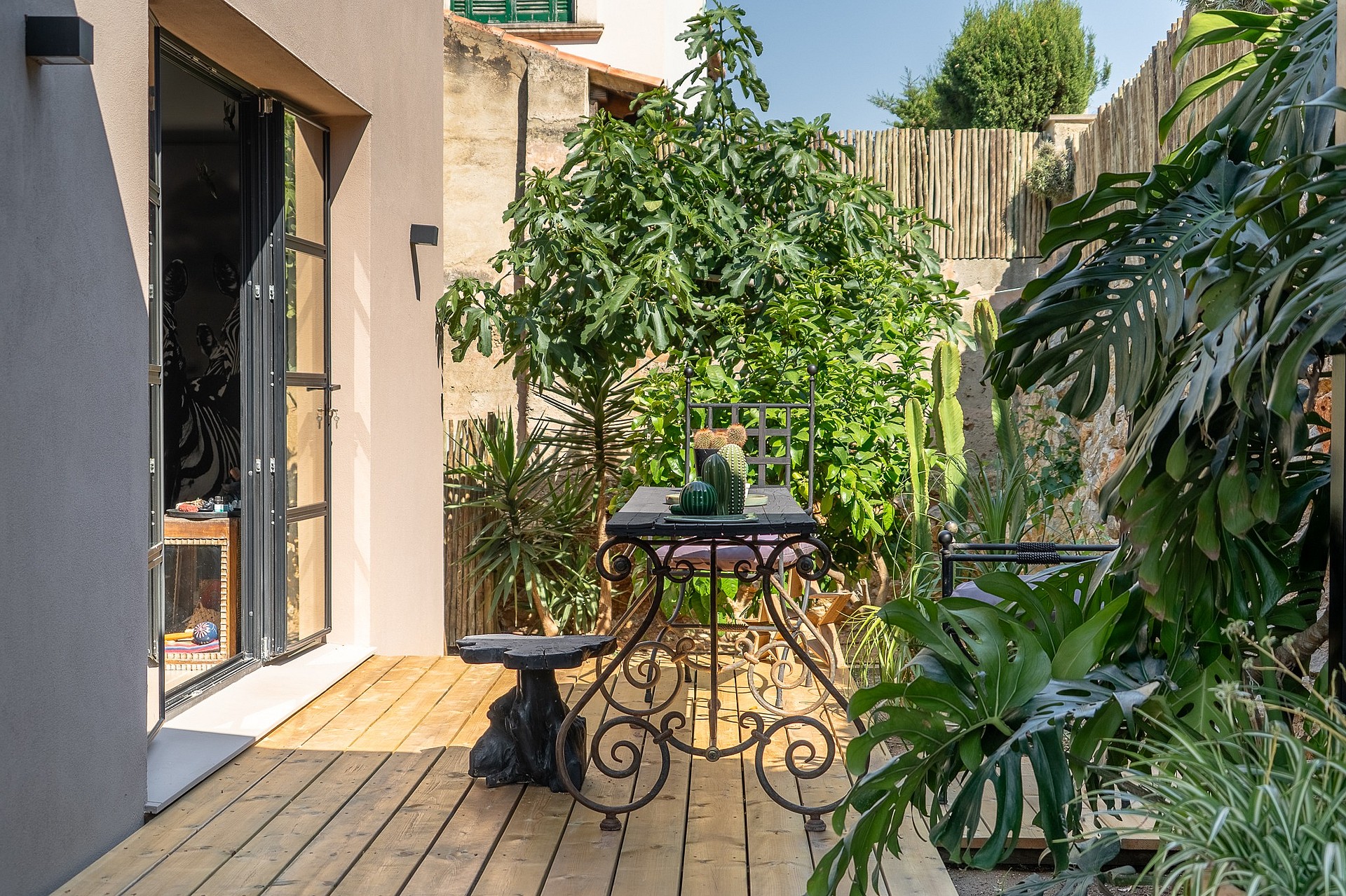 A small outdoor garden space with light-colored wood decking, a wrought iron table, and various potted green plants.