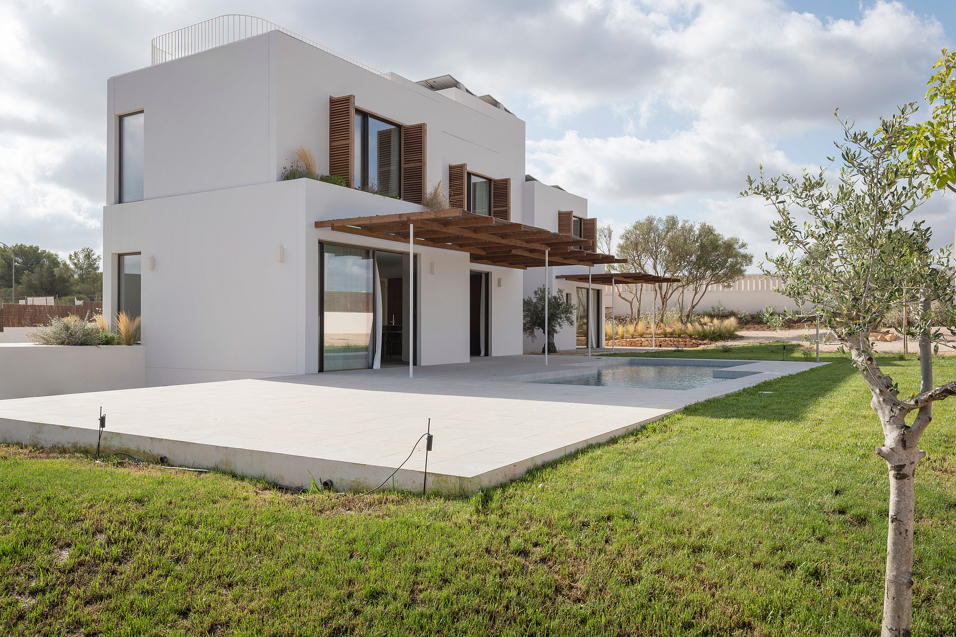 Modern two-story white villa with wooden shutters, a large pergola, and stone terrace overlooking a green lawn.