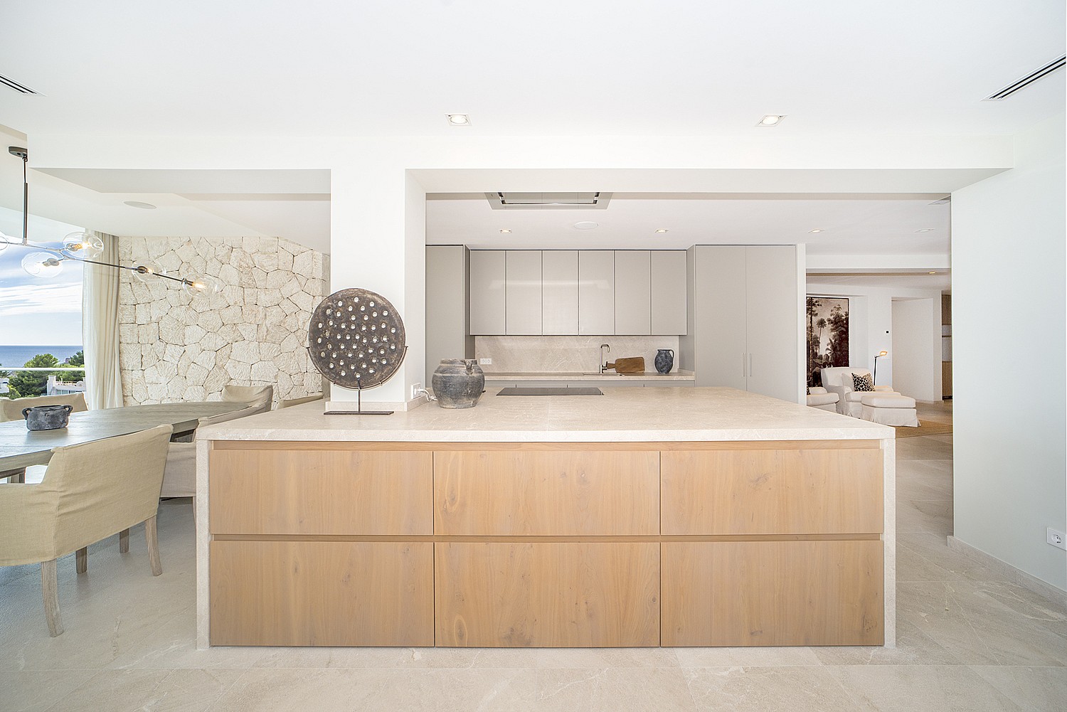 Modern open-plan kitchen featuring a large wooden island with a stone countertop and light wood cabinets, set against a white wall.