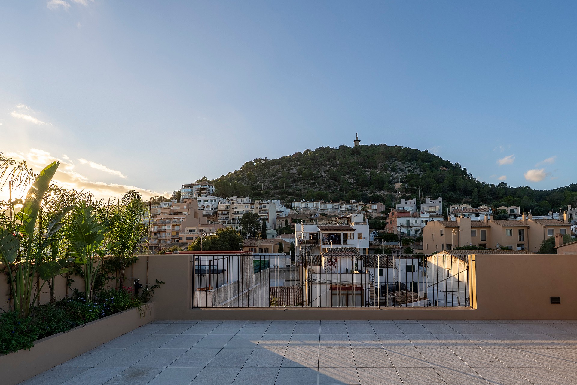 Stone-paved rooftop terrace with iron railings and planters, facing a hillside village and forested mountain peaks.