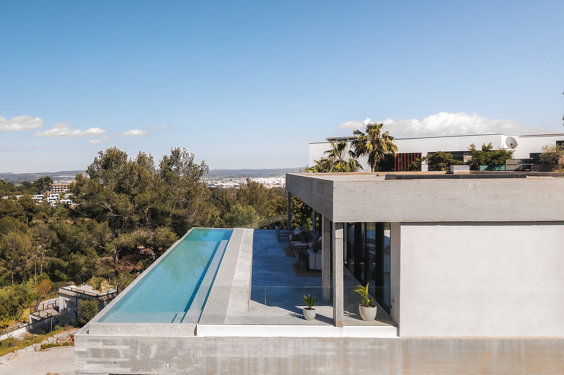 Elevated view of a long infinity lap pool on a grey stone terrace of a minimalist concrete villa overlooking the valley.