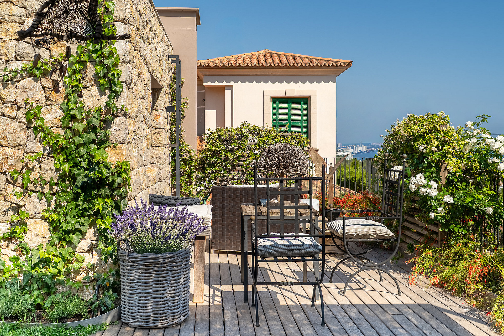 A sunny stone-walled terrace in Genova featuring metal garden furniture, a lavender planter, and climbing ivy plants.
