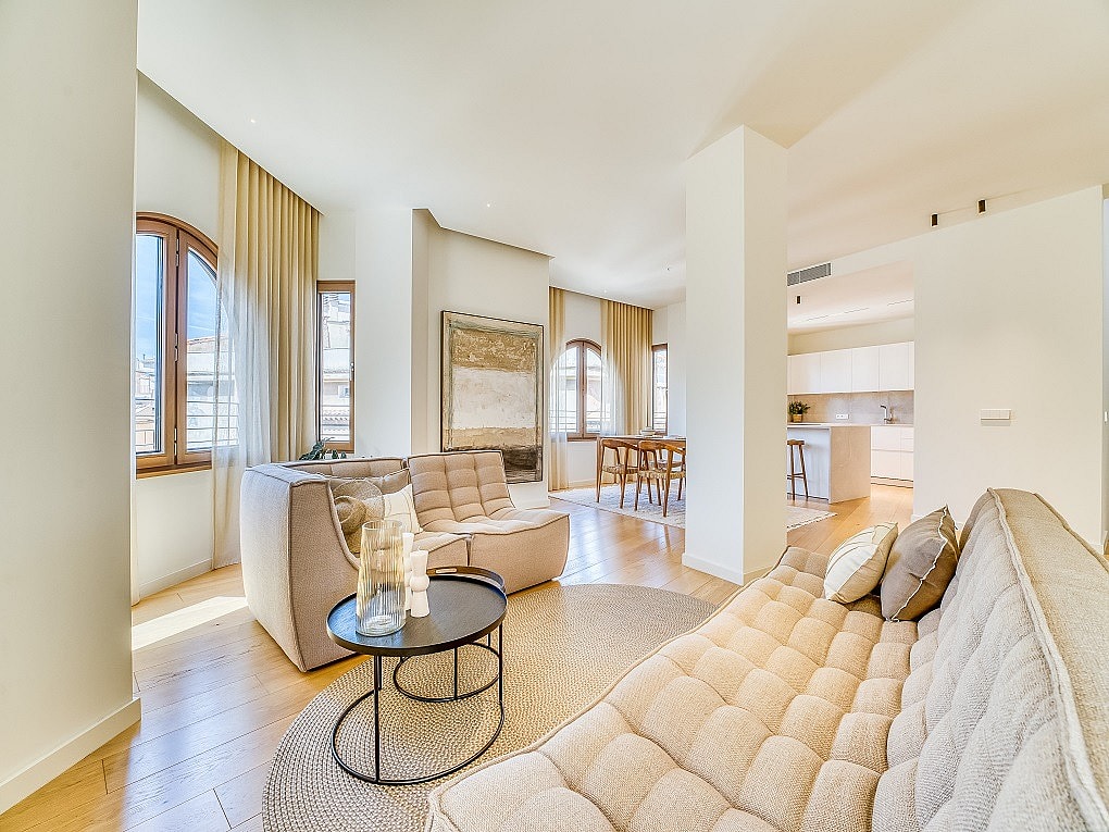 A bright living room in a penthouse with arched windows, light oak flooring, beige modular sofas, and a dining area in the background.