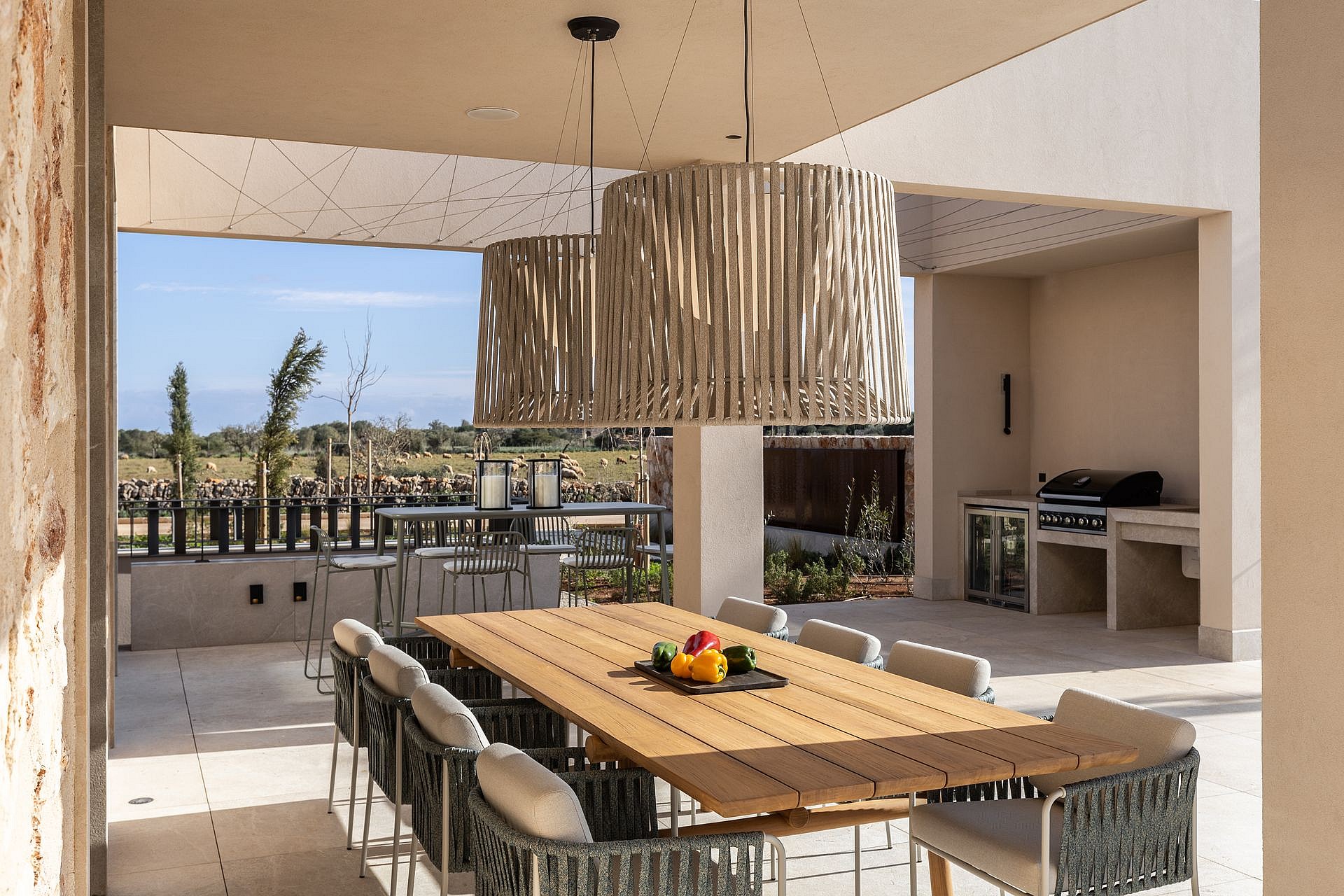 A spacious outdoor dining patio features a long wooden table, woven chairs, and a stone-built summer kitchen, looking towards mountain fields.