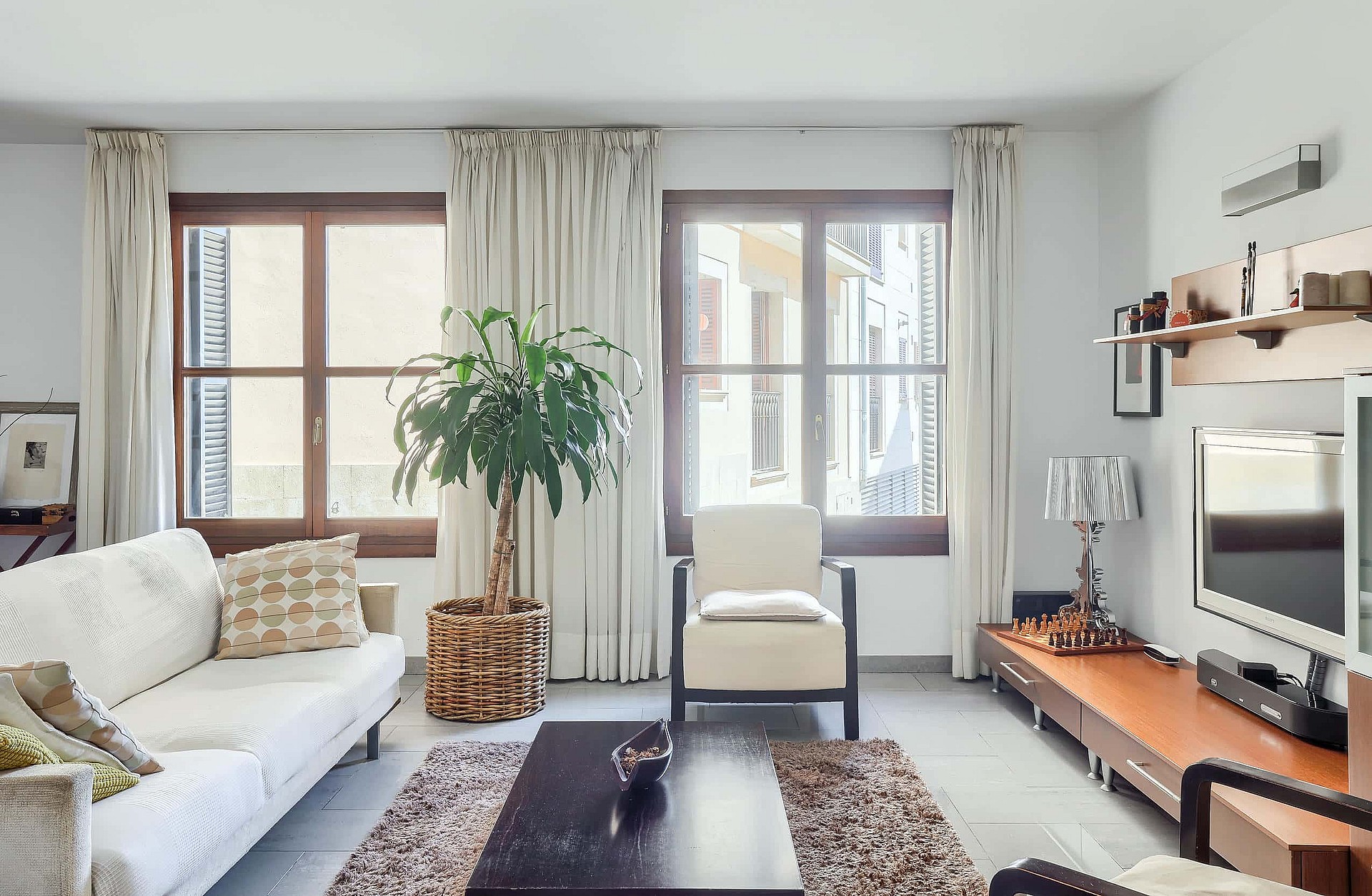 A living room featuring a white sofa, cream armchair, wooden media console, and a tall indoor plant in a woven basket.