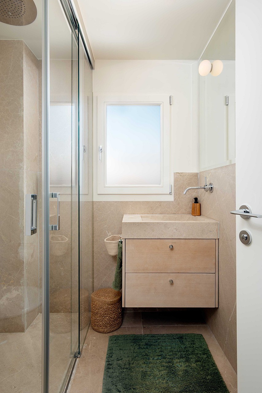 A modern small bathroom featuring a floating wooden vanity, a glass-enclosed walk-in shower, and beige stone wall tiling.