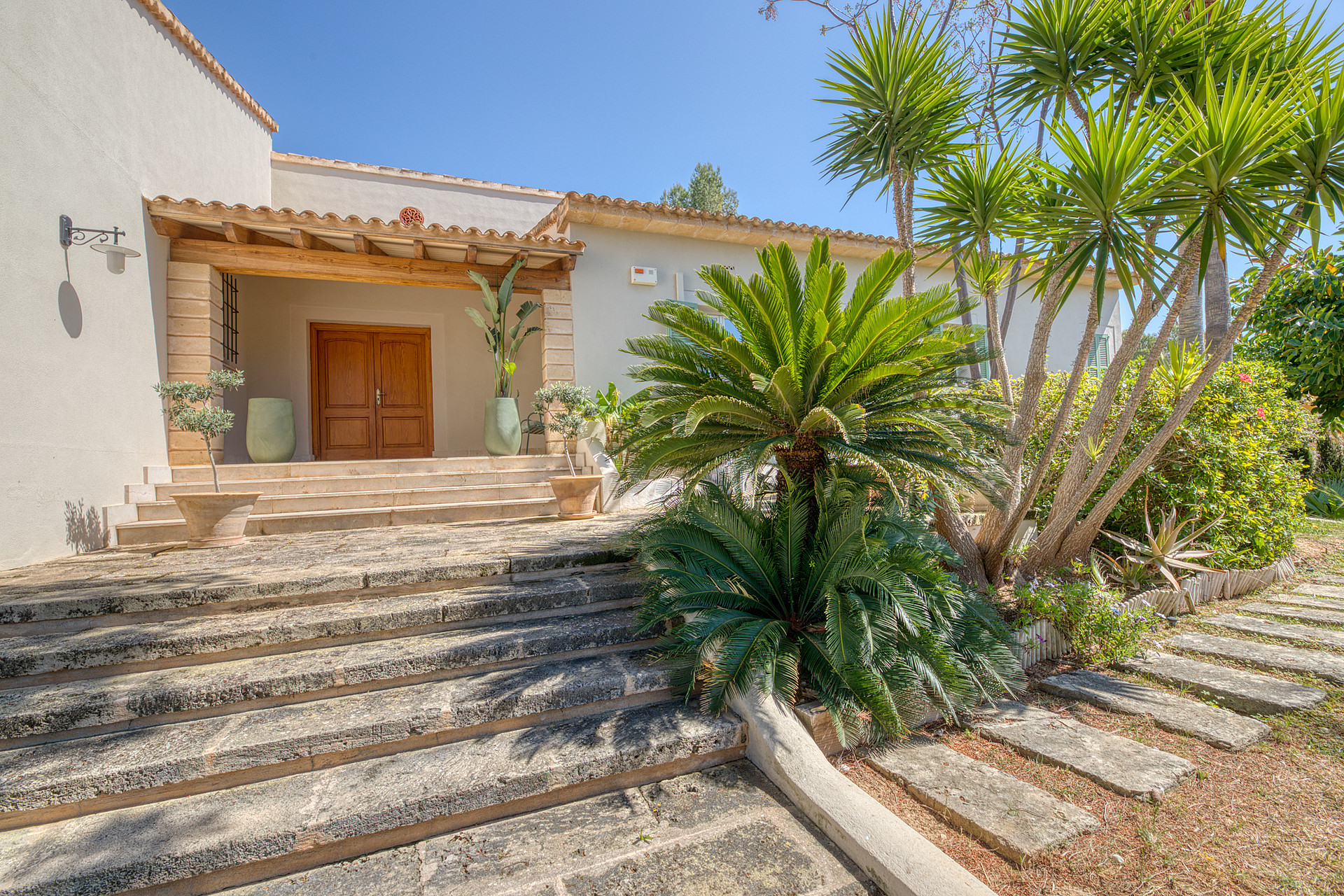 A wide stone staircase leads up to a wooden-framed entrance door under a terracotta-tiled porch, flanked by palm trees.
