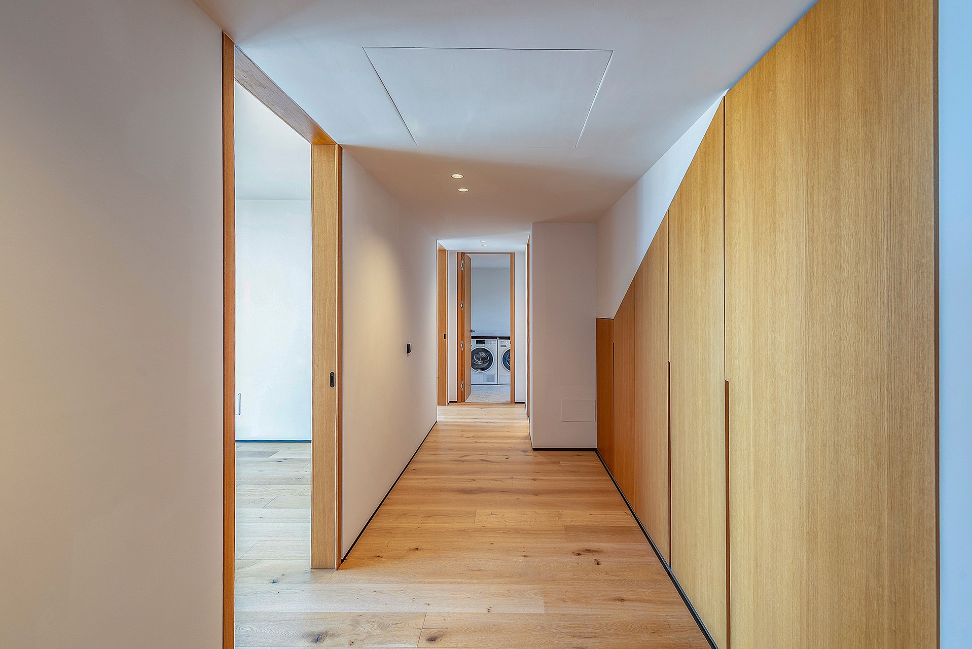 A modern residential hallway with light oak wood floors and integrated floor-to-ceiling oak wood cabinetry on the right wall.
