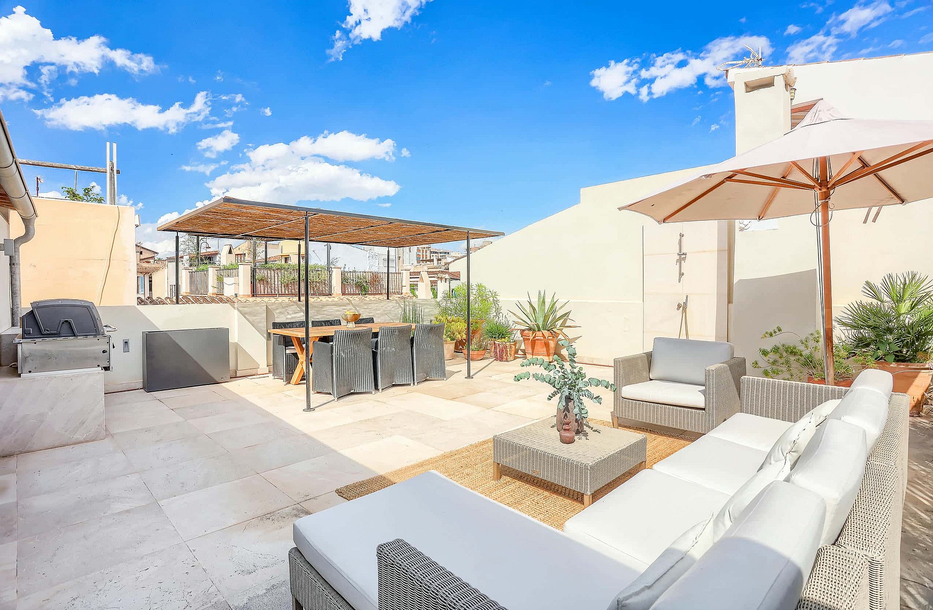 A bright rooftop terrace with a dining area under a wooden pergola and a lounge seating area with white cushions and a large cream umbrella under a clear blue sky.