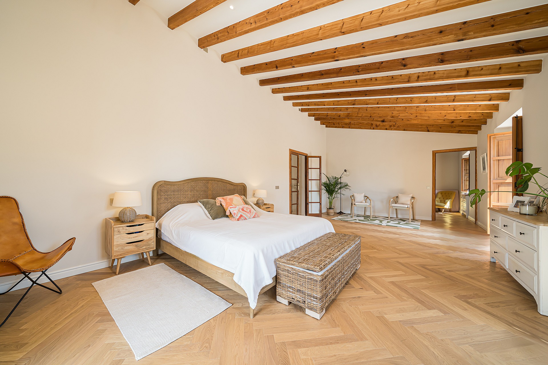 A spacious master bedroom featuring light oak herringbone floors, exposed ceiling beams, and woven furniture accents.