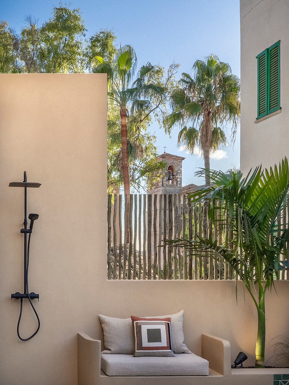 Minimalist outdoor shower with black fixtures next to a built-in bench, framed by palm trees and a village tower view.
