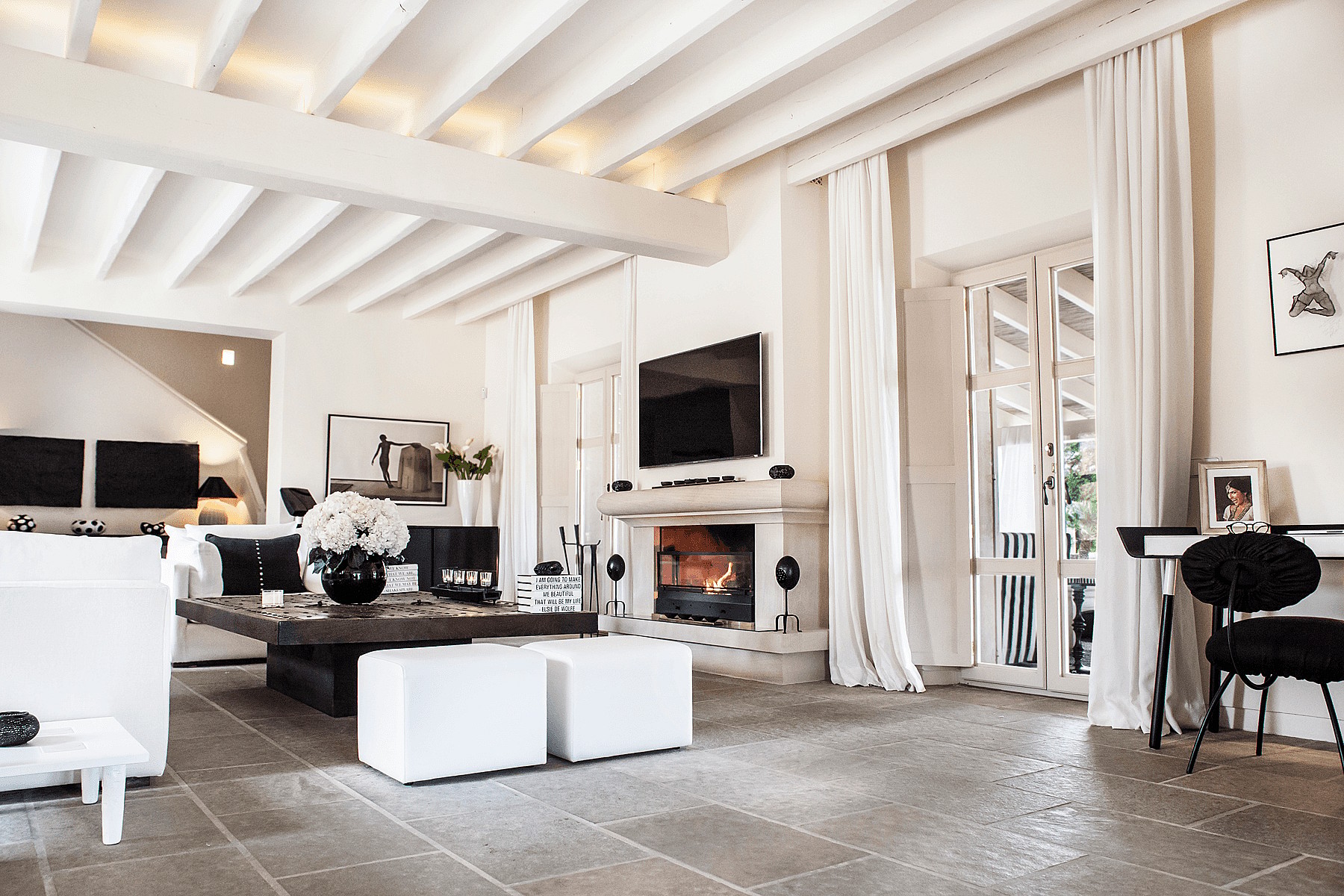 Bright living area with a stone fireplace, exposed beam ceiling, stone flooring, and white linen curtains.