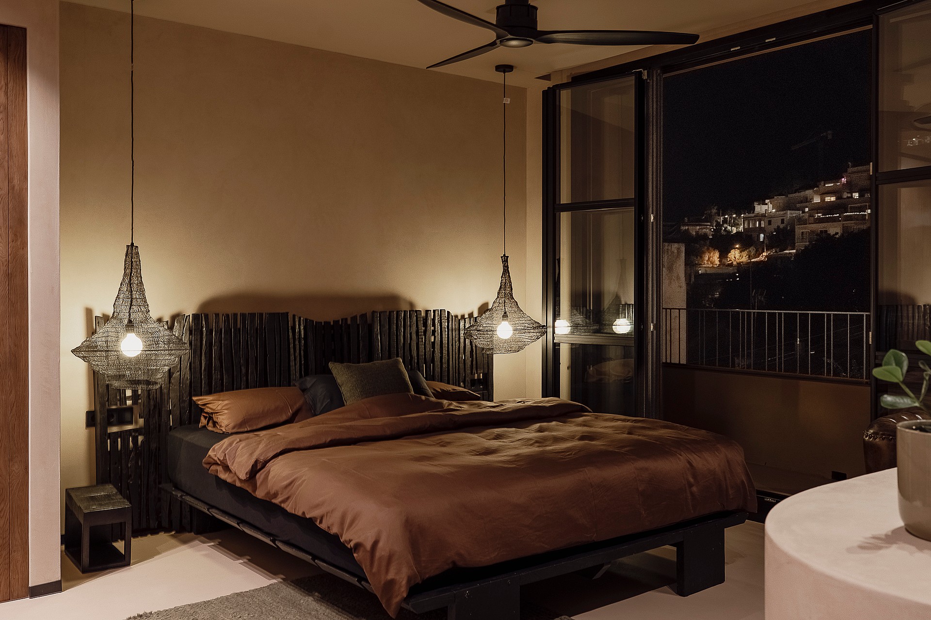 Bedroom with a queen-sized bed, dark wood headboard, and two decorative pendant lights. Large sliding glass door reveals a night view of hills.