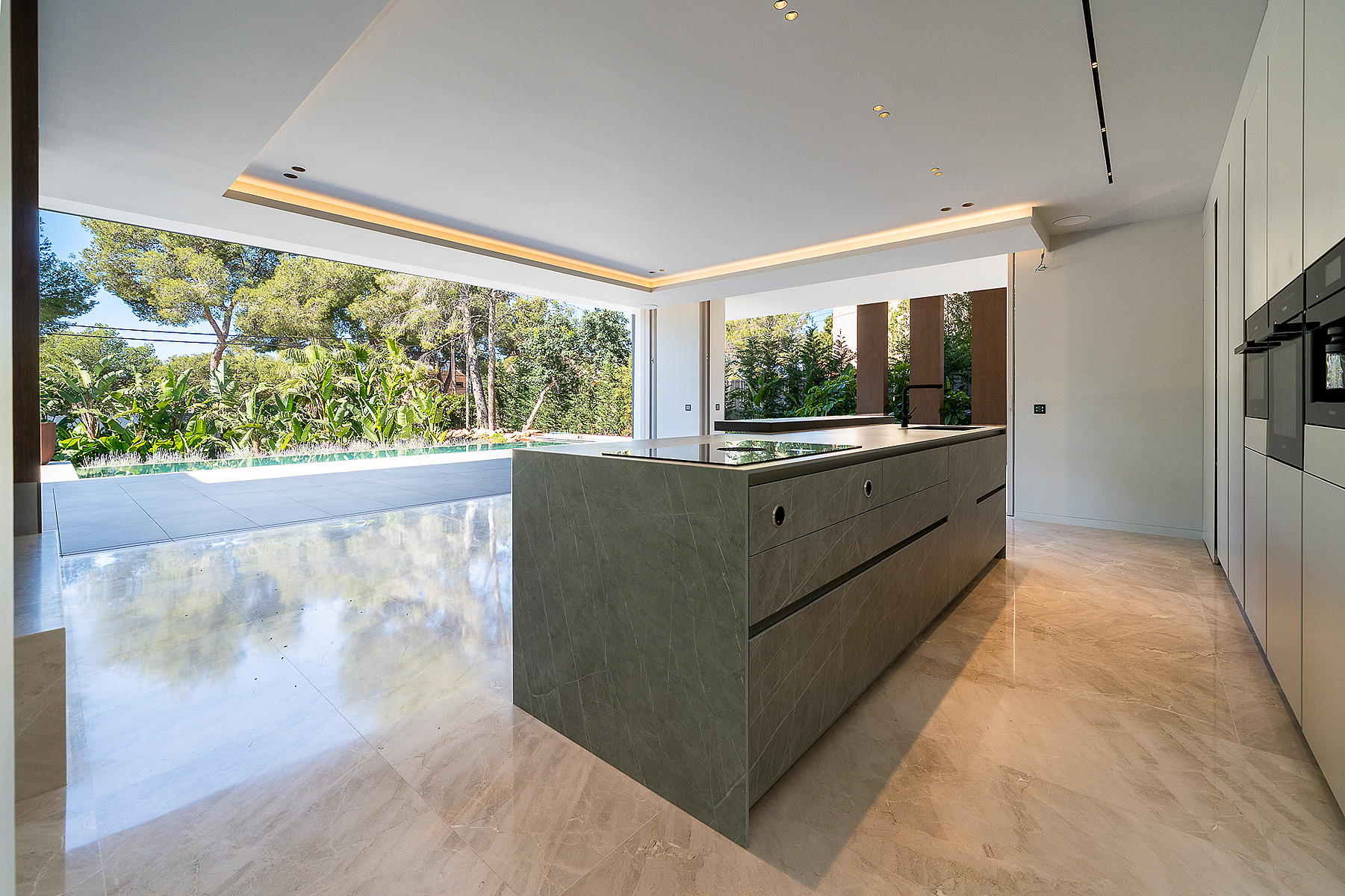 Modern kitchen with a large dark stone island, pale marble floors, and wide sliding doors opening to a garden terrace.