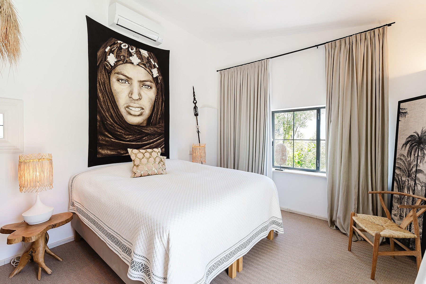 A bedroom featuring a bed with a white quilt, a large decorative wall tapestry above the headboard, beige floor-to-ceiling curtains, and a natural wood side table.