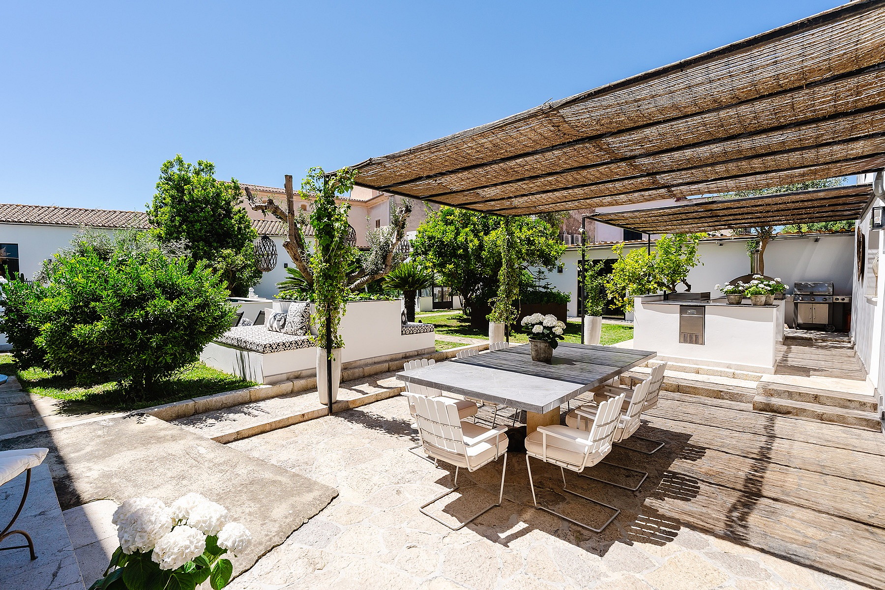 A sunny outdoor stone terrace featuring a dining table under a woven reed pergola, surrounded by lush green garden plants.