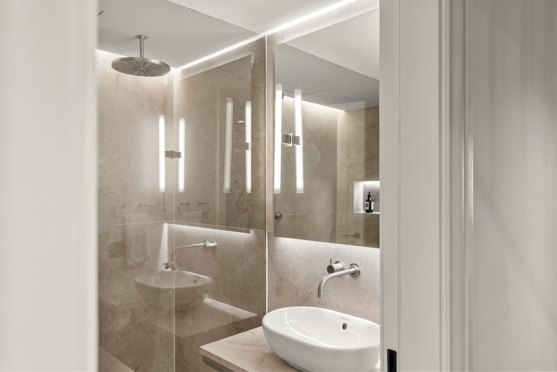 Modern bathroom featuring marble walls, a glass walk-in shower with rainfall head, and mirrors with integrated lighting.