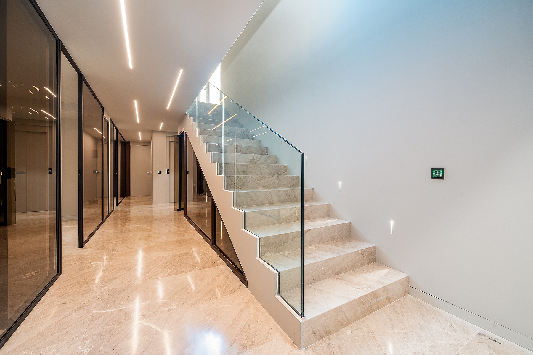 A wide interior hallway with a beige marble staircase featuring a frameless glass railing and recessed LED ceiling lighting.