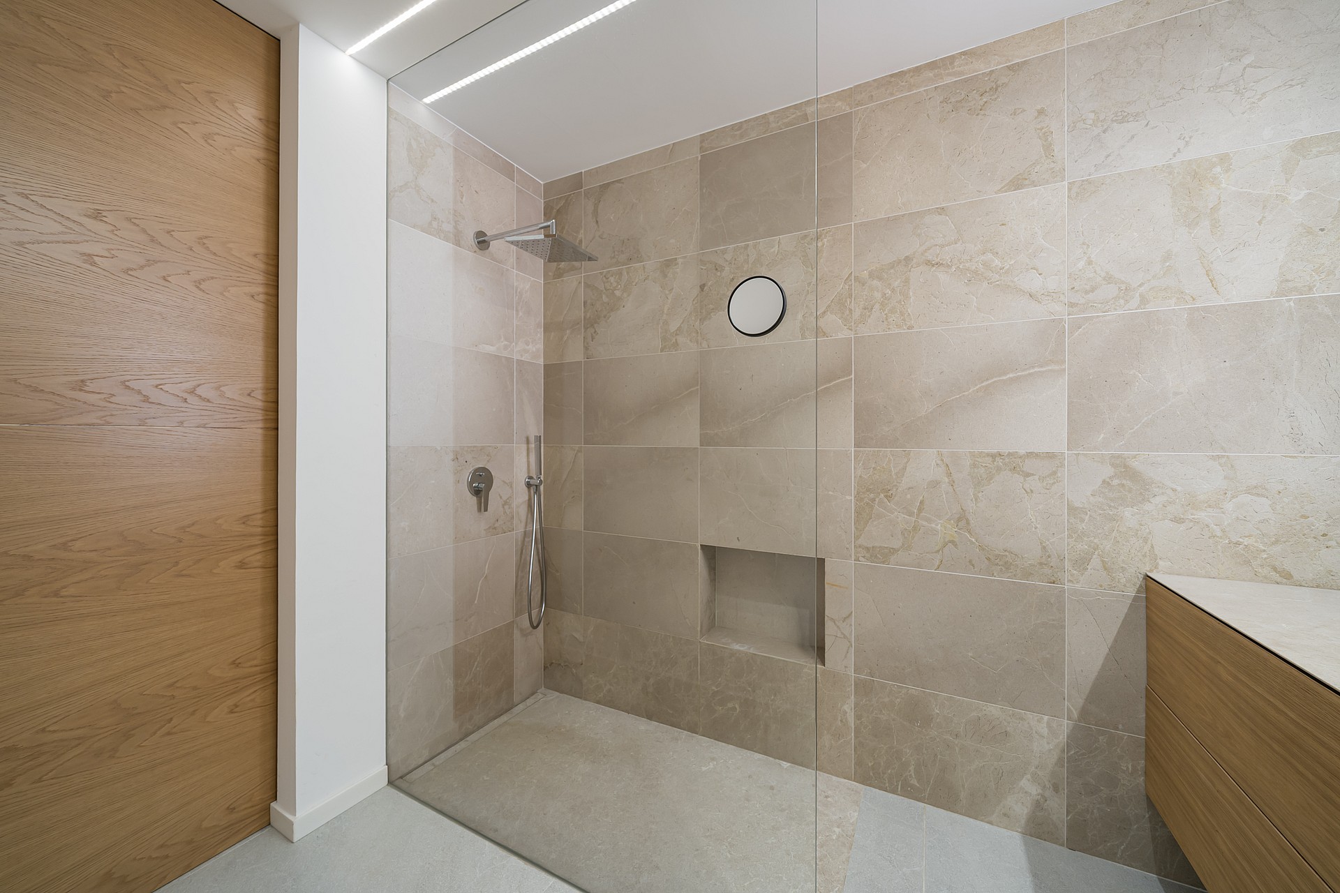 A walk-in shower with large-format beige stone tiles, a glass partition, and integrated recessed shelving.