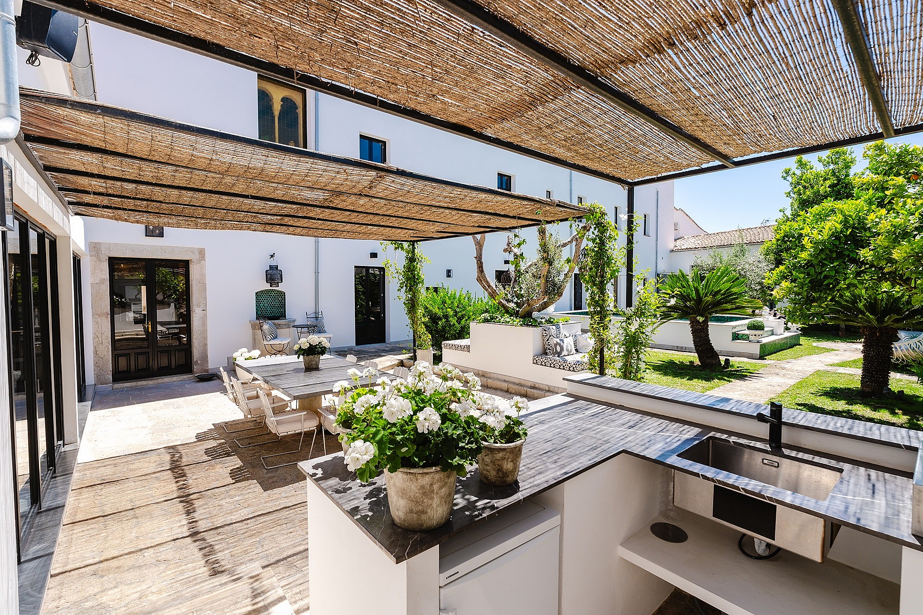 An outdoor kitchen with stone counters and a sink sits under a woven reed canopy, adjoining a paved terrace and garden.