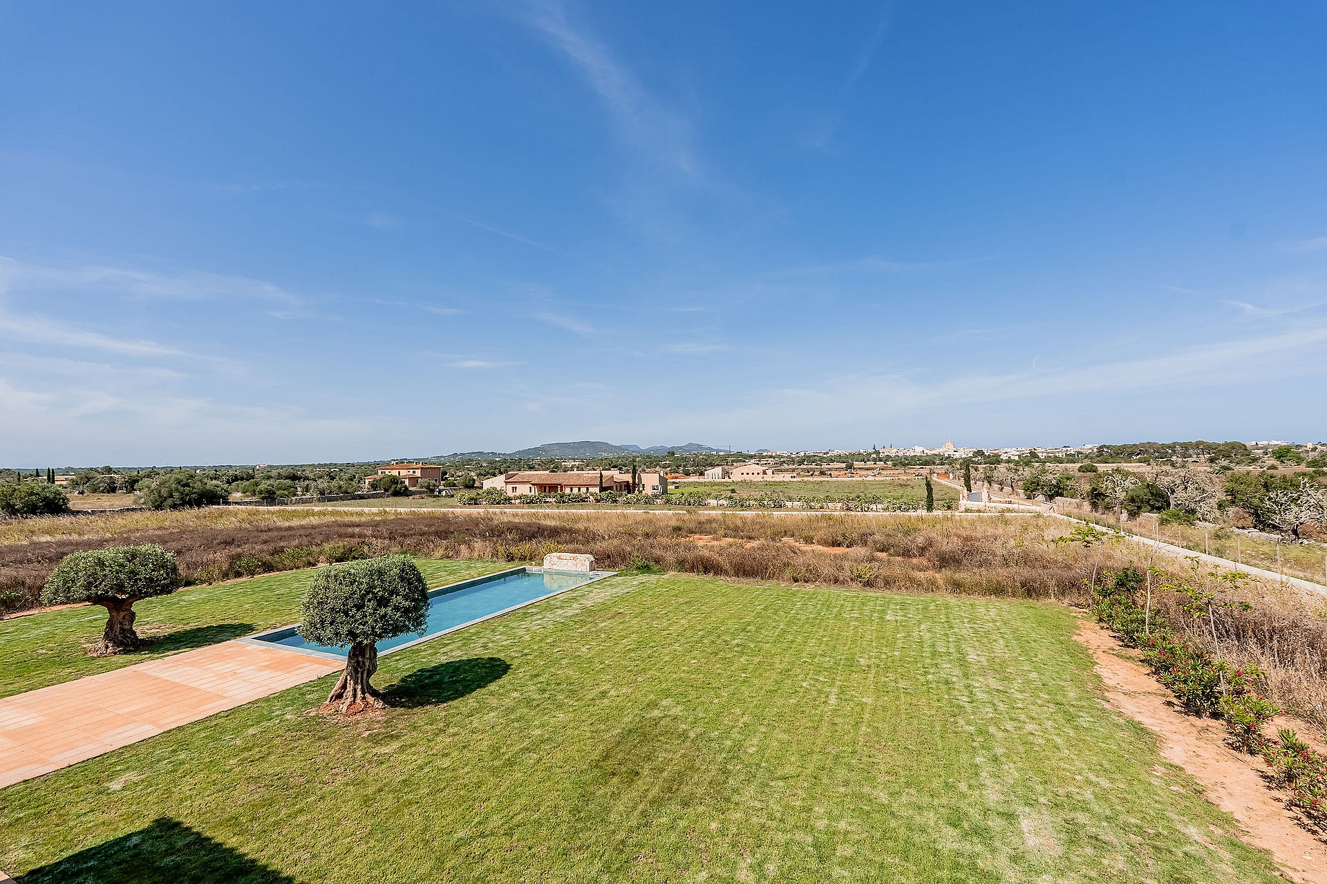 An elevated view over a green lawn and rectangular pool toward distant mountains in the Santanyí countryside.