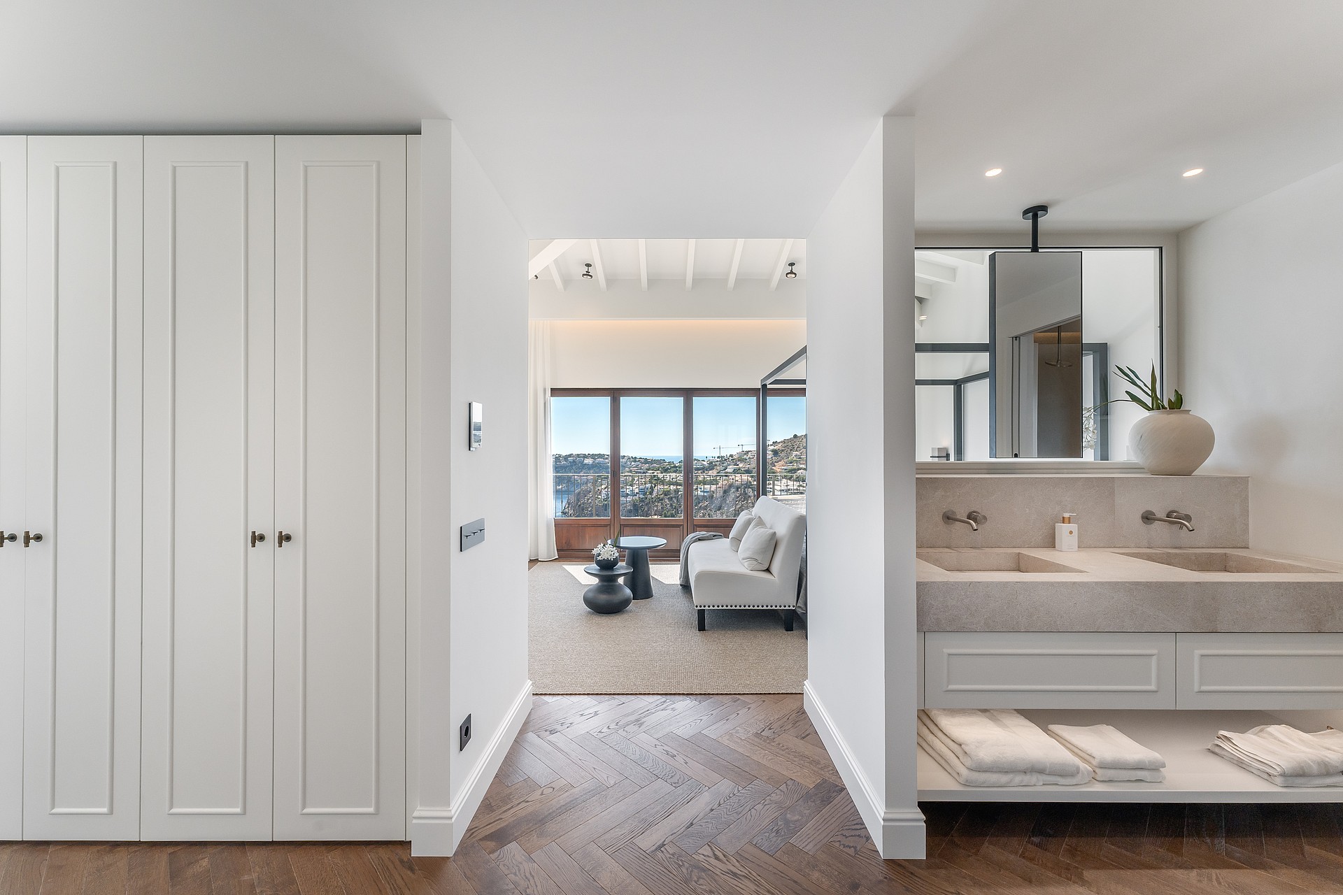 En-suite bathroom with double stone sinks and herringbone floors leading into a bedroom with a sofa and sea views.