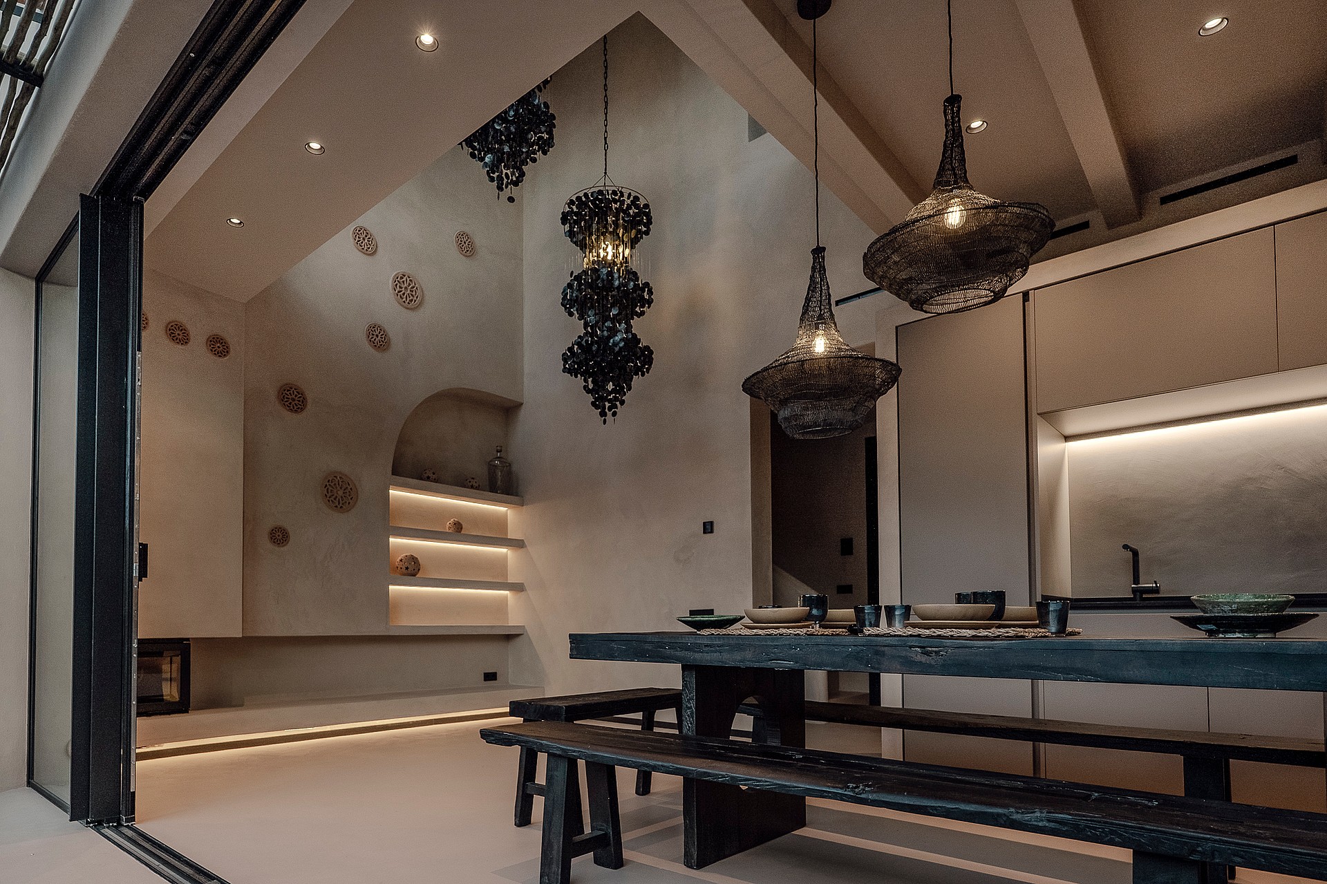 Dining area featuring a dark wooden farmhouse table and benches, sculptural black pendant lights, and arched wall niches with soft integrated illumination.